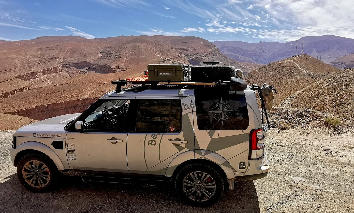 A white SUV with rooftop gear parked on a dirt trail in a remote desert canyon landscape with red rock formations and mountains in the distance.