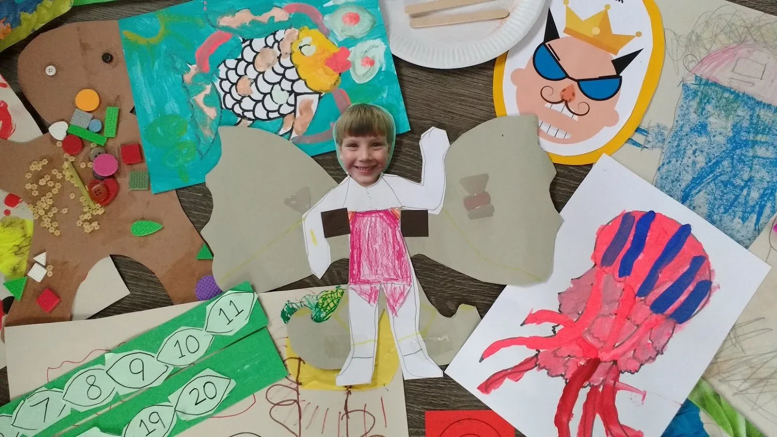 A colorful craft project featuring a child's photo as the face of a butterfly with paper wings, surrounded by various drawings and arts and crafts materials, including a crab illustration and a prize ribbon chart.