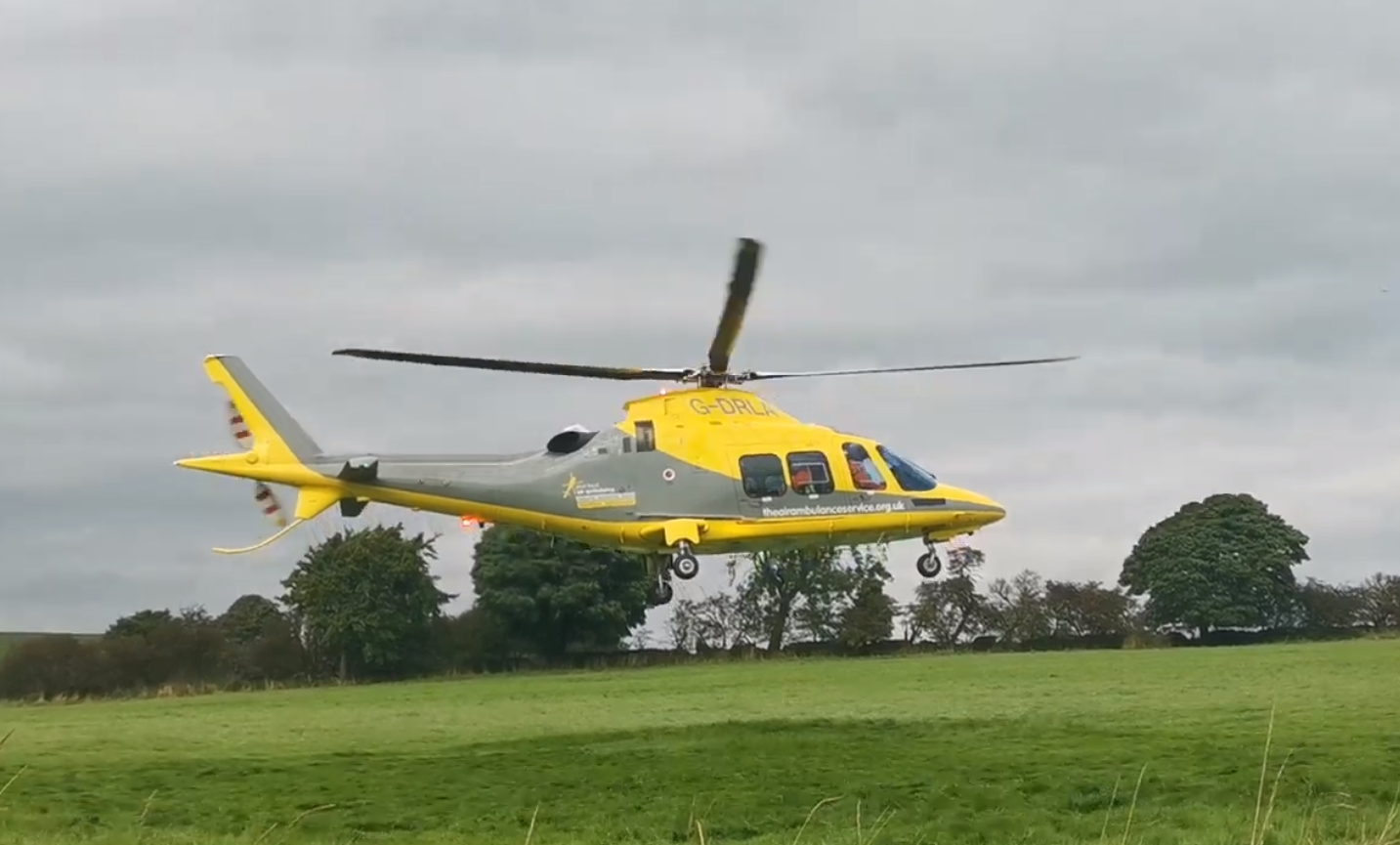 Yellow and gray helicopter flying low over a grassy field with trees and cloudy sky in the background.