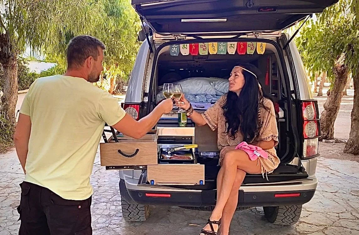 A man and woman enjoying drinks during a picnic in the back of an open SUV with trees in the background.