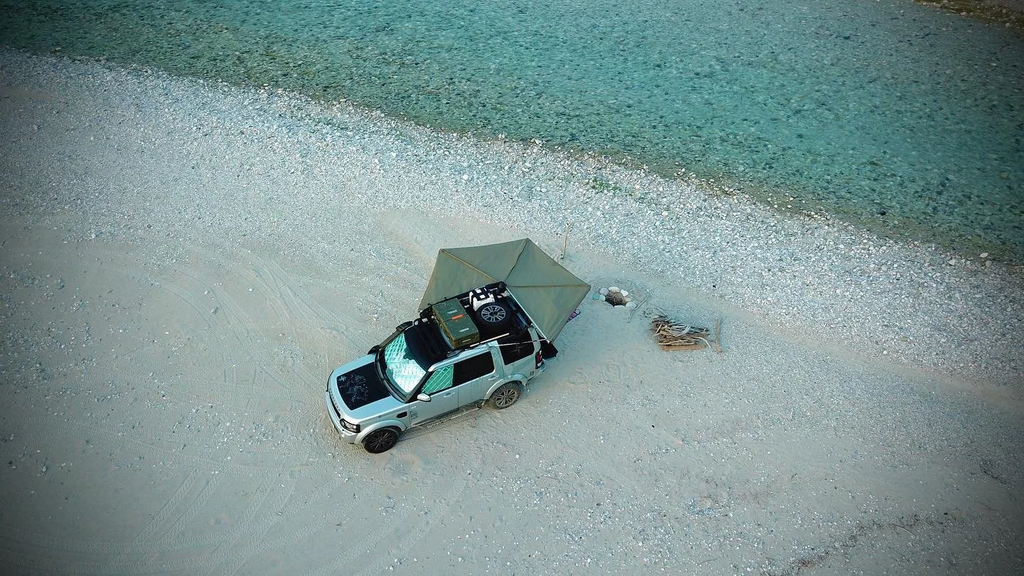 A silver off-road vehicle parked on sandy terrain near a body of water, with a tent set up on the roof and camping supplies nearby.