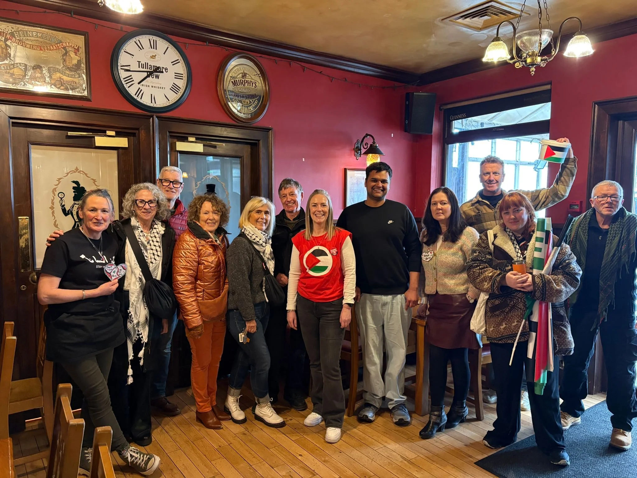 A group of people posing together inside a pub or restaurant, with red walls, wooden doors, and clocks showing 9:13 and 9:09. Some are smiling and holding party accessories. There is a window with a Guinness sign and colorful decorations.