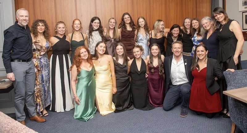 Group of  camogie female players and coaches including Mark Barry dressed in formal attire, posing together indoors in front of a wooden wall, celebrating their Junior A win 2025.