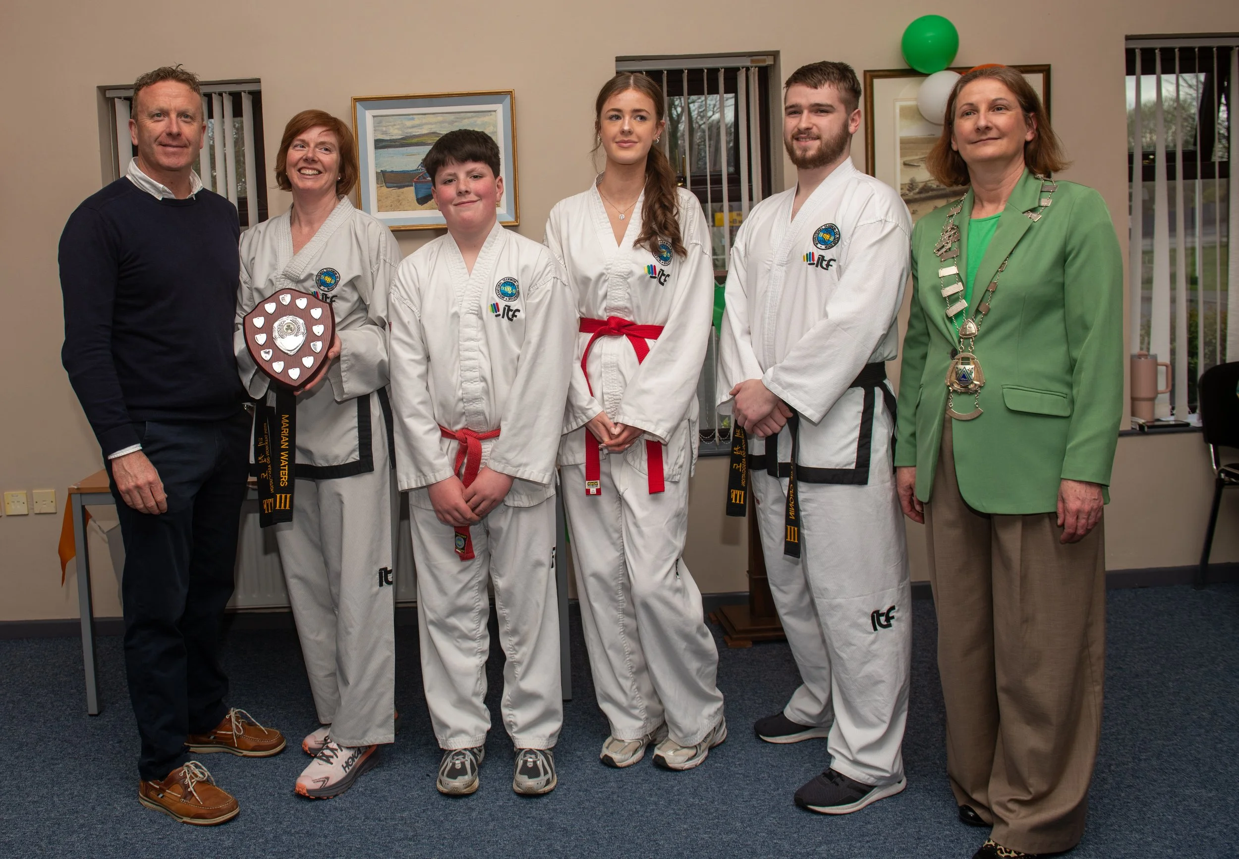 Group of seven people standing indoors, five are in martial arts uniforms, and two are in casual attire, with one holding a shield-shaped award, behind them on the wall are landscape paintings and balloons.
