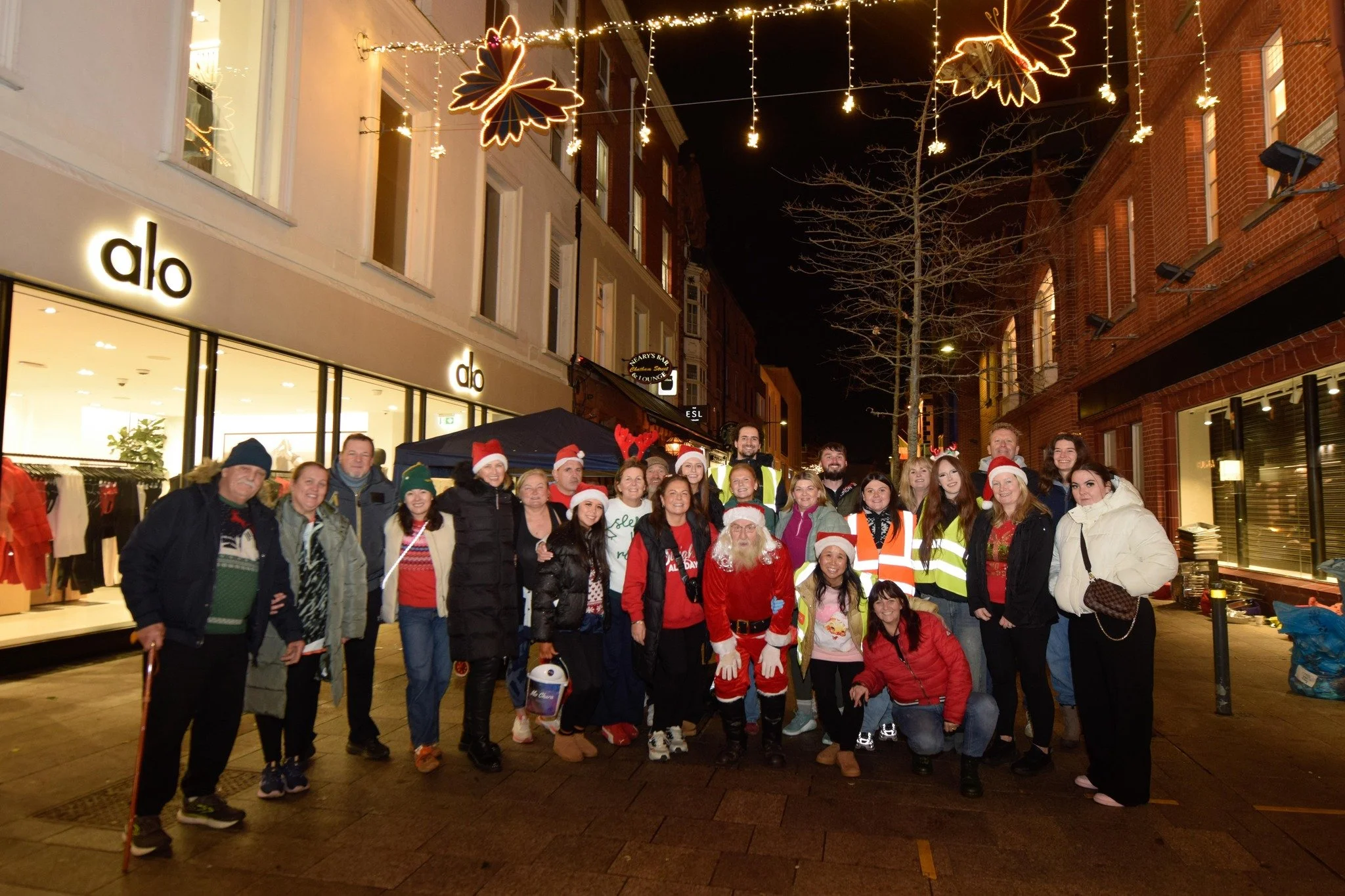 A group of people dressed in Christmas attire and winter clothing gathered outdoors at night, with festive lights and Christmas decorations overhead, including large illuminated butterfly-shaped lights and star-shaped lights hanging from above.