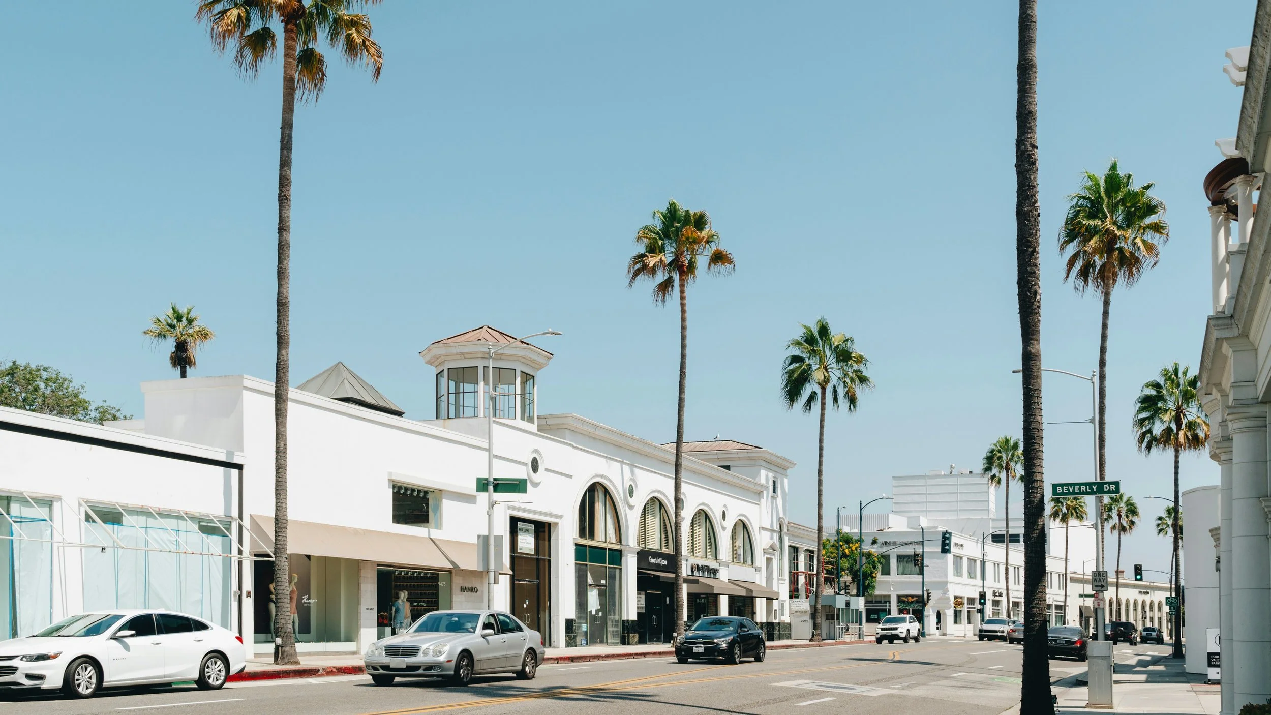 A Los Angeles, California street scene with white buildings, tall palm trees, parked cars, and a street sign that reads Beverly Dr, in a sunny location with a clear blue sky.