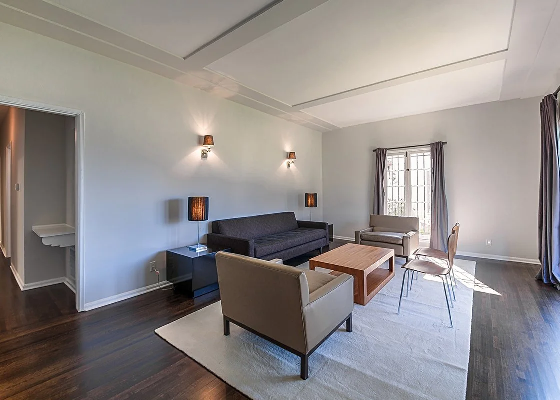 Living room with white walls, dark wood floor, gray sofa, beige armchairs, wooden coffee table, dark side tables with black lamps, and large windows with dark curtains.