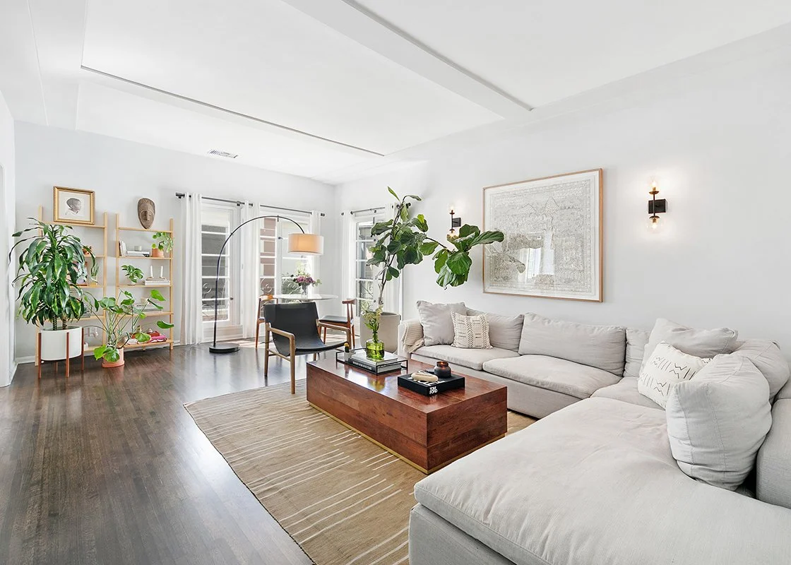 A modern living room with white walls, dark hardwood floors, a large white sectional sofa, a wooden coffee table, a black armchair, a tall green houseplant, a beige rug, and large windows with white curtains.