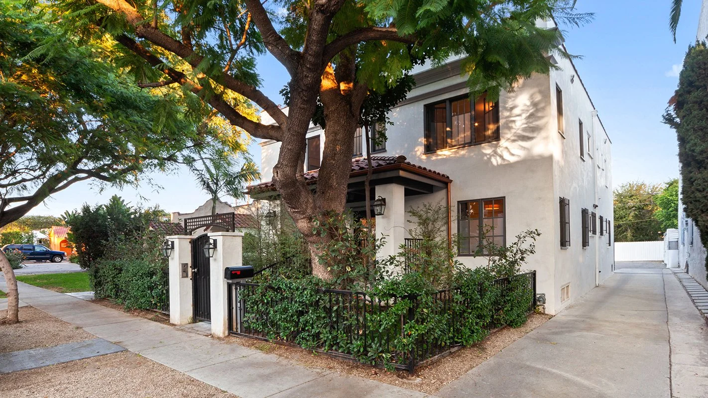 A white three-story house with black window grilles, surrounded by greenery and trees. There is a sidewalk, a black gate, and a small enclosed yard with bushes.