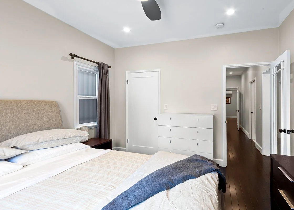 A bedroom with a bed, nightstand, window with curtain, dresser, and hallway with hardwood floors.