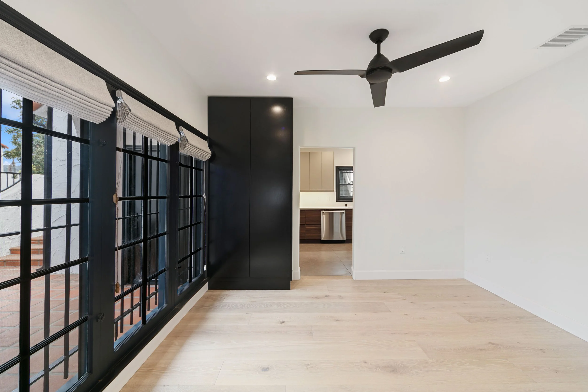 Empty room with hardwood floors, large black-framed windows with blinds, black corner cabinet, ceiling fan, viewed into a kitchen area with dark wood cabinets and stainless steel dishwasher.