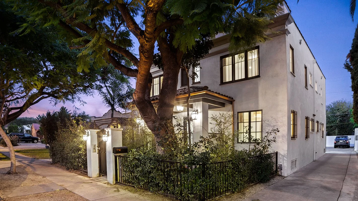 A two-story white house with black window frames, surrounded by a small garden with shrubbery and a fence. There are trees in the front yard, and the house is lit up in the evening.