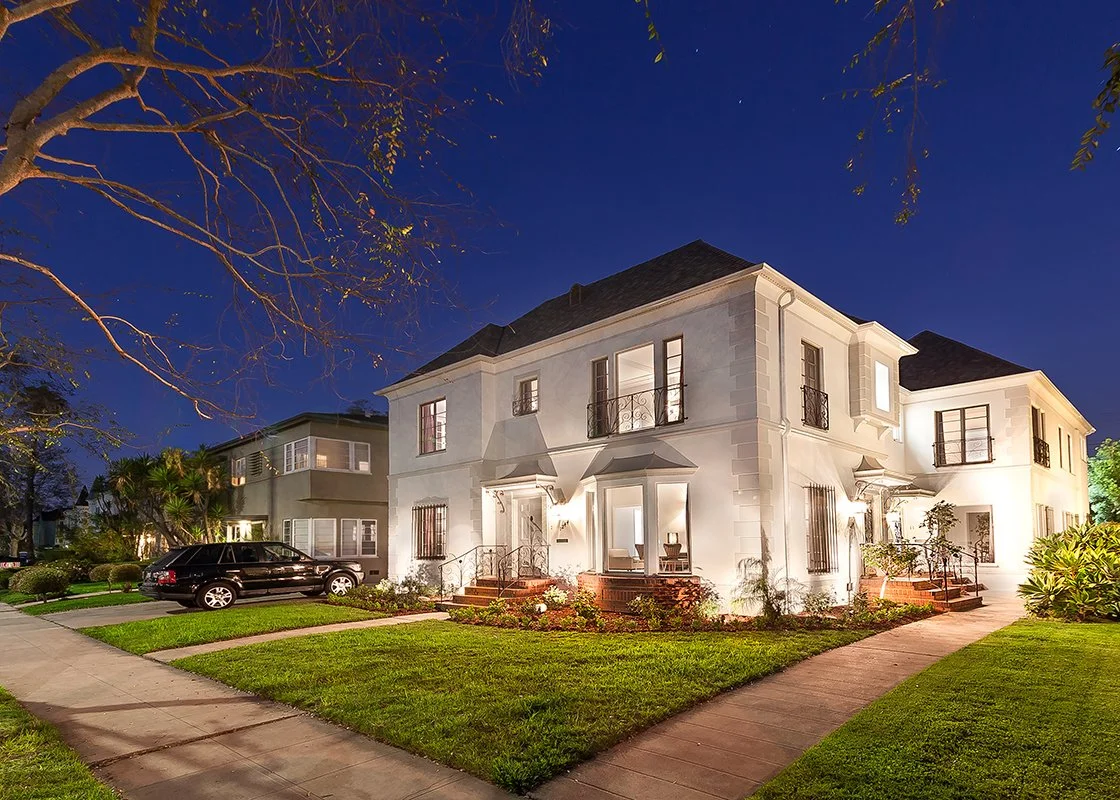 A well-lit two-story house with a manicured lawn and a sidewalk in front, viewed at night. There are trees and shrubs around the house, and a black vehicle is parked on the street.
