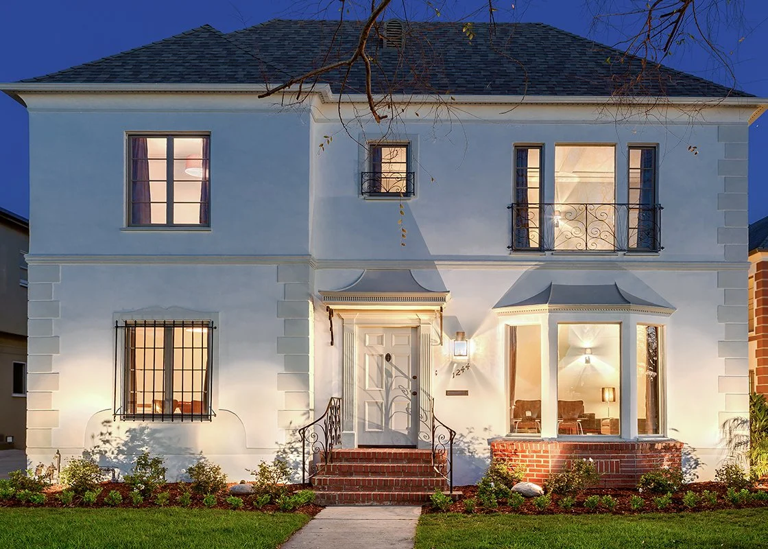 Front view of a two-story white house with brick steps, a dark roof, and lit windows at dusk.