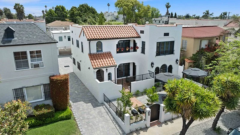 A white, two-story house with a red-tiled roof, surrounded by a small yard with outdoor seating, umbrellas, and a gated entrance, in a neighborhood with similar style houses and lush greenery.