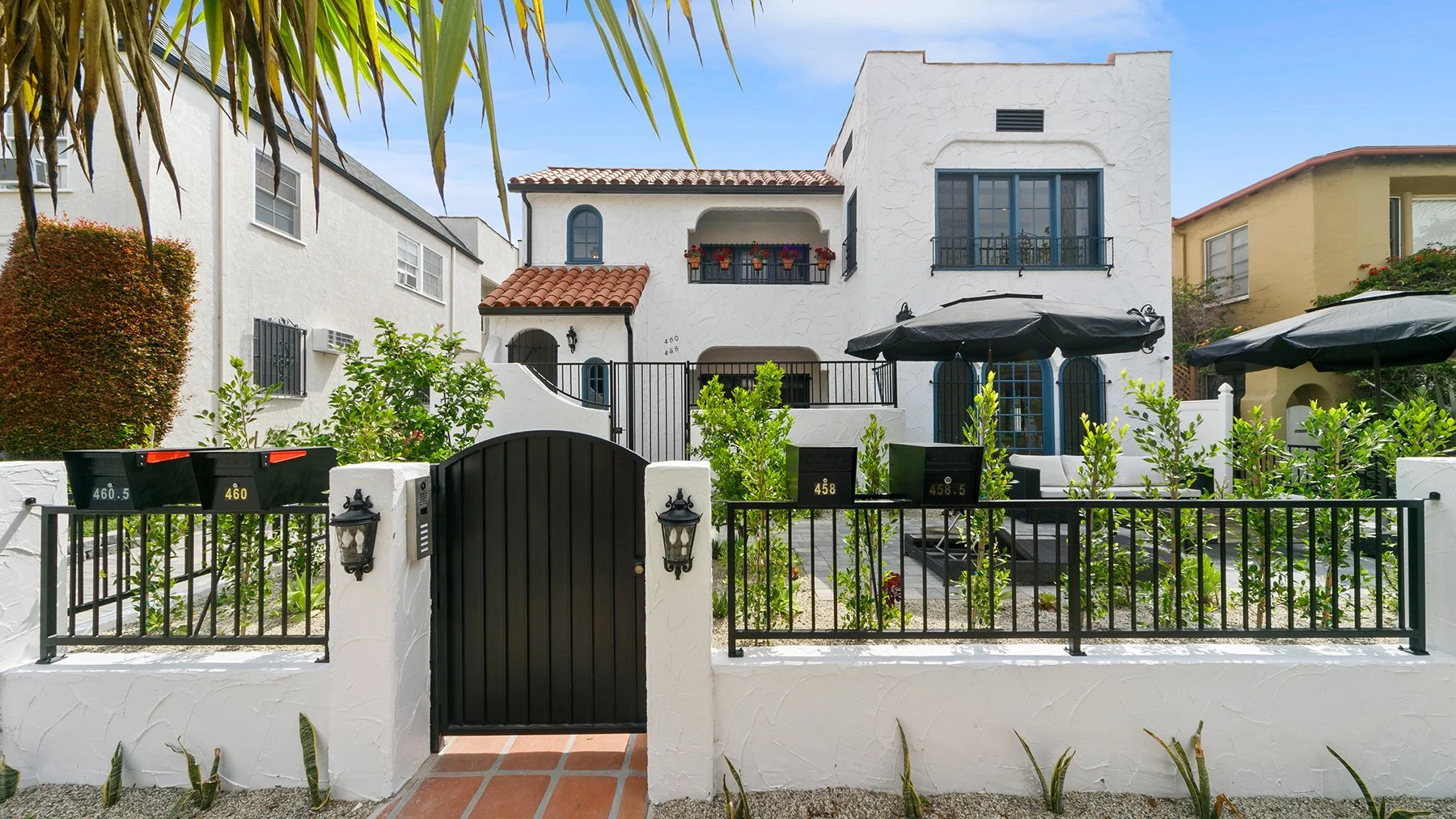 White Mediterranean-style house with black gates, outdoor seating, umbrellas, and lush greenery in front yard.