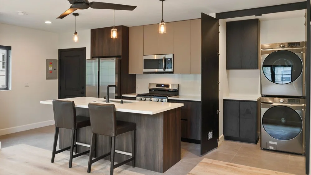 Modern kitchen with dark wood cabinets, stainless steel appliances, and stacked washer and dryer. Includes a kitchen island with two black chairs and hanging pendant lights.
