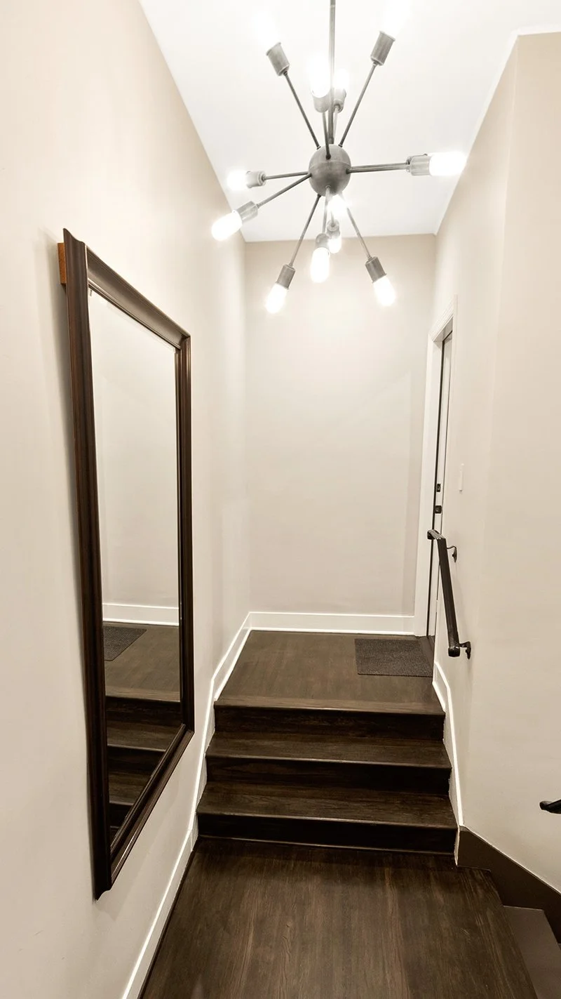 Interior view of a staircase landing with dark wood steps, a mirror on the left wall, a door with a handrail on the right, and a modern chandelier overhead.