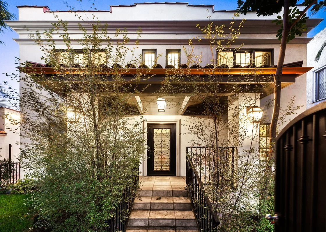 Front door of a two-story house with a small staircase, surrounded by bushes, with black wrought-iron fencing, exterior lighting, and a tree on the right.
