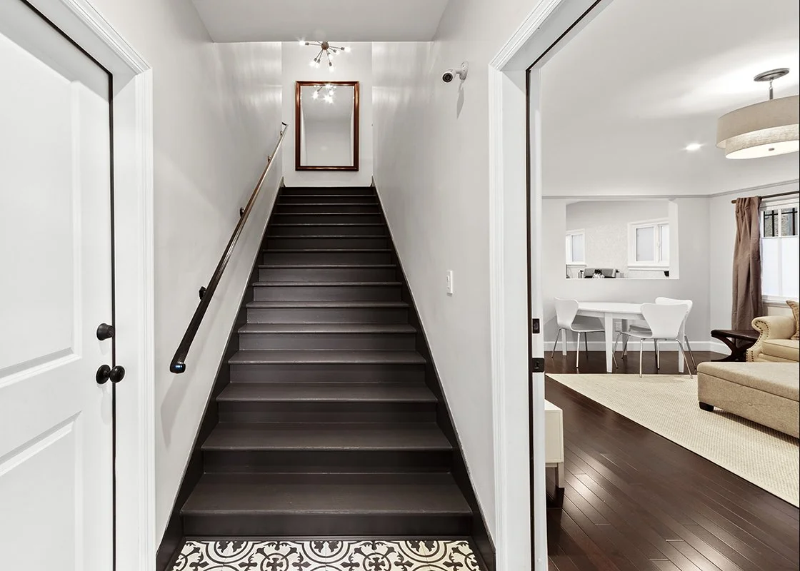 Interior view of a home featuring a staircase, a mirror at the top, and part of a living room with hardwood floors, a beige sofa, and a dining area with white chairs and a table.