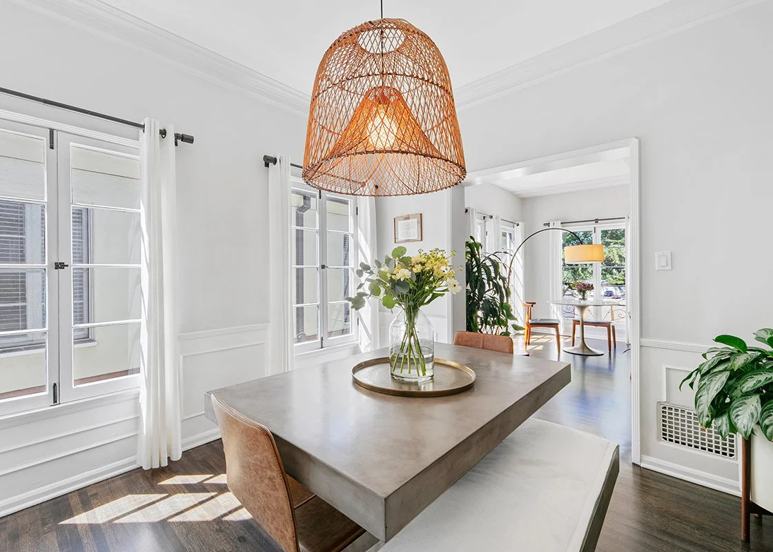 A bright dining room with large windows, a concrete table with a glass vase of white and yellow flowers, a woven pendant light, and green plants in the background.