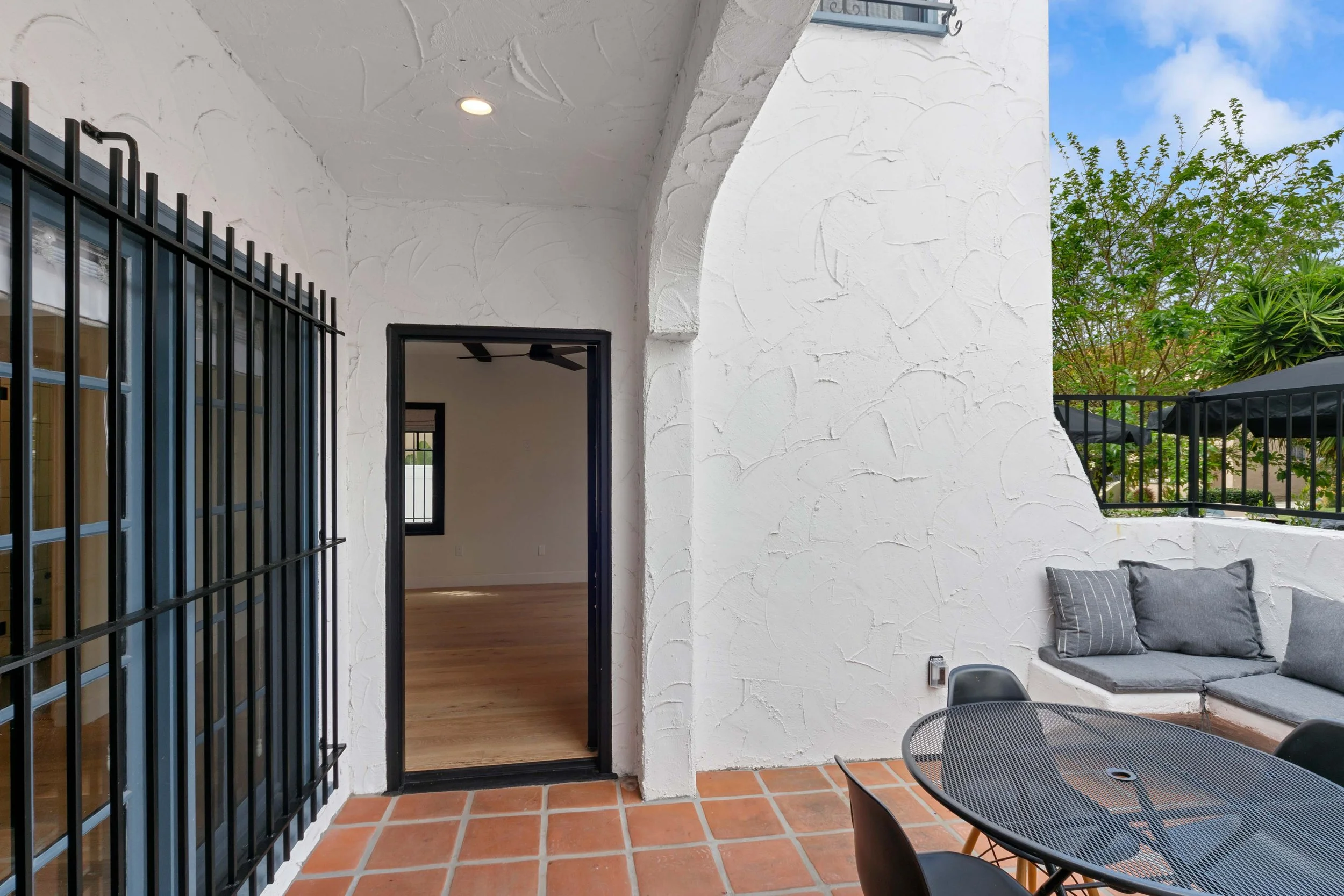 Outdoor patio area with terracotta tile flooring, white stucco walls, black metal door frame, window with metal bars, built-in seating with gray cushions, black outdoor table, and black chairs, with lush green trees and blue sky in the background.