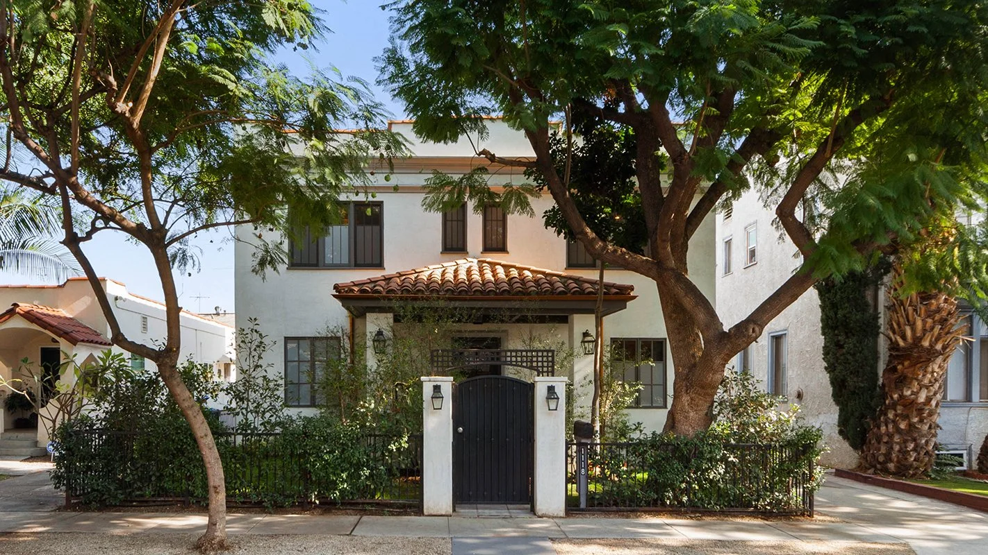 A white two-story house with a tiled roof, dark window frames, and a gated entrance, surrounded by trees and a low fence, on a sunny day.