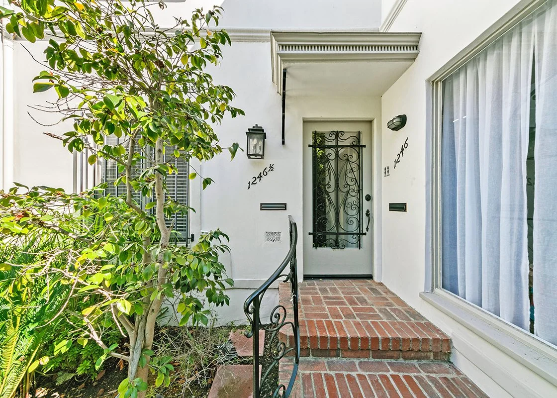 Front porch of a house with white exterior walls, a black wrought iron door, brick steps, a large window with curtains, green leafy plants to the left, and house numbers 12462 and 12464.