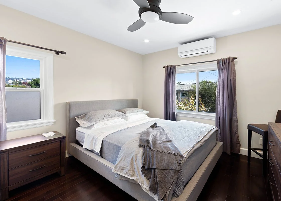 A bedroom with a grey bed, dark wood nightstand, and dresser. There are two windows with purple curtains, an air conditioning unit, and a ceiling fan.