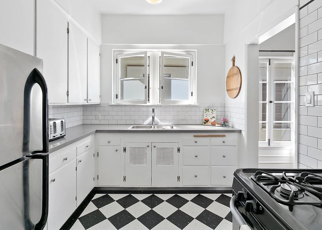 Bright, white kitchen with black and white checkered floor, white cabinets, a window over the sink, and a black gas stove in the foreground.