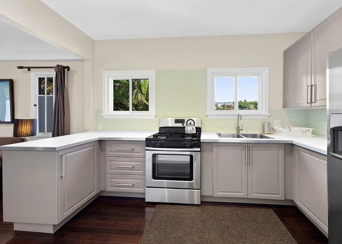 Modern kitchen with white cabinets, gray countertops, stainless steel stove and fridge, two windows, and a brown floor mat.
