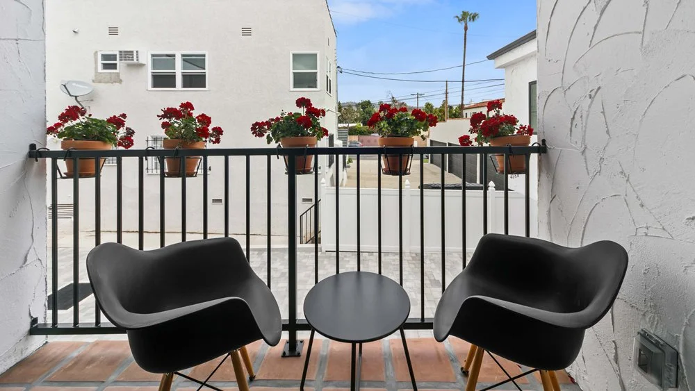 A small balcony with two black modern chairs and a small black table, overlooking a courtyard with flower pots on a railing and a background of buildings and palm trees.