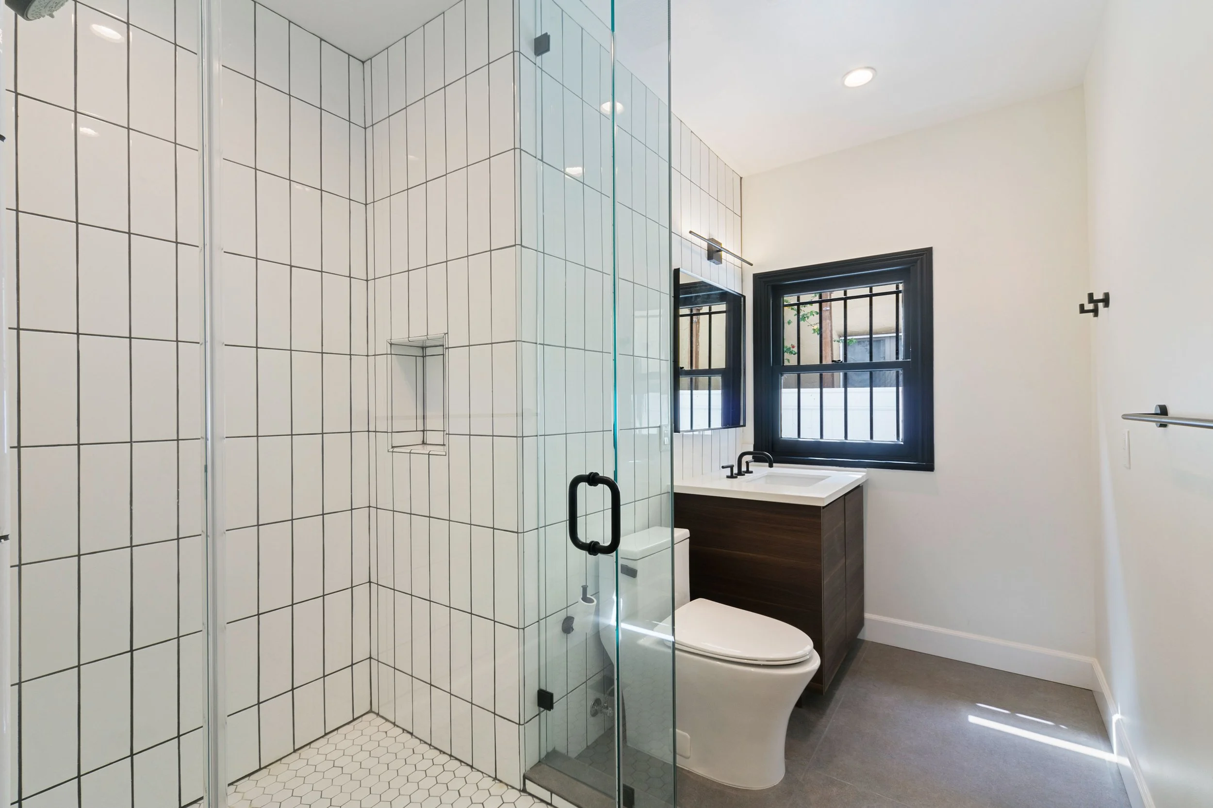 Modern bathroom with a glass-enclosed shower, white tile walls, a toilet, a dark wooden vanity with a white sink, black fixtures, and a window with black trim.