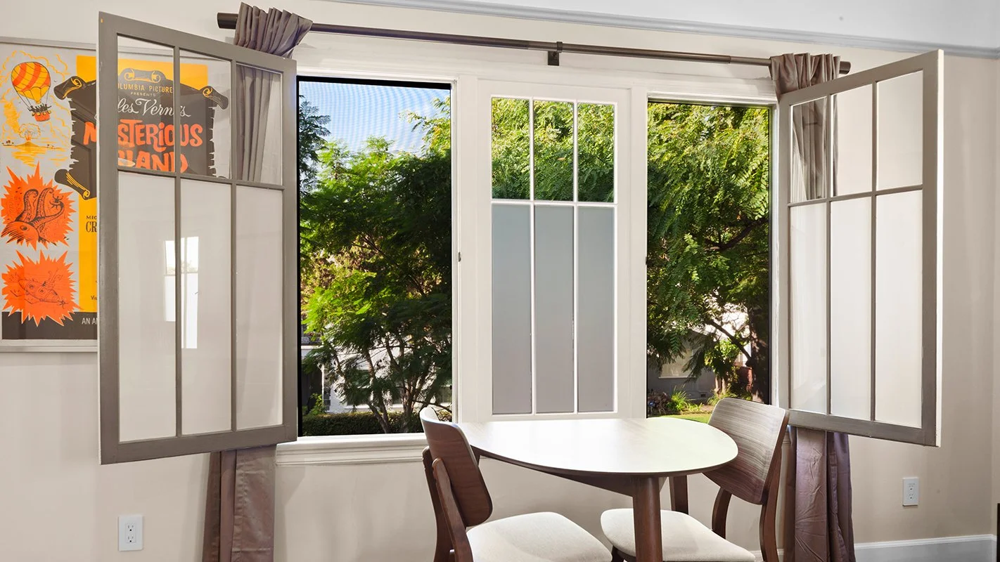 Open window with a view of green trees outside, a small table, and chairs inside a room.