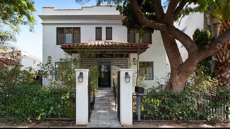 A white two-story house with a tiled roof, black window frames, and a gated front entrance surrounded by greenery and trees.
