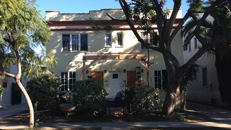 A two-story building with a cream-colored facade, surrounded by trees and shrubbery. Two people are standing near a bench in front of the building. The scene is sunny with shadows cast on the building and street.