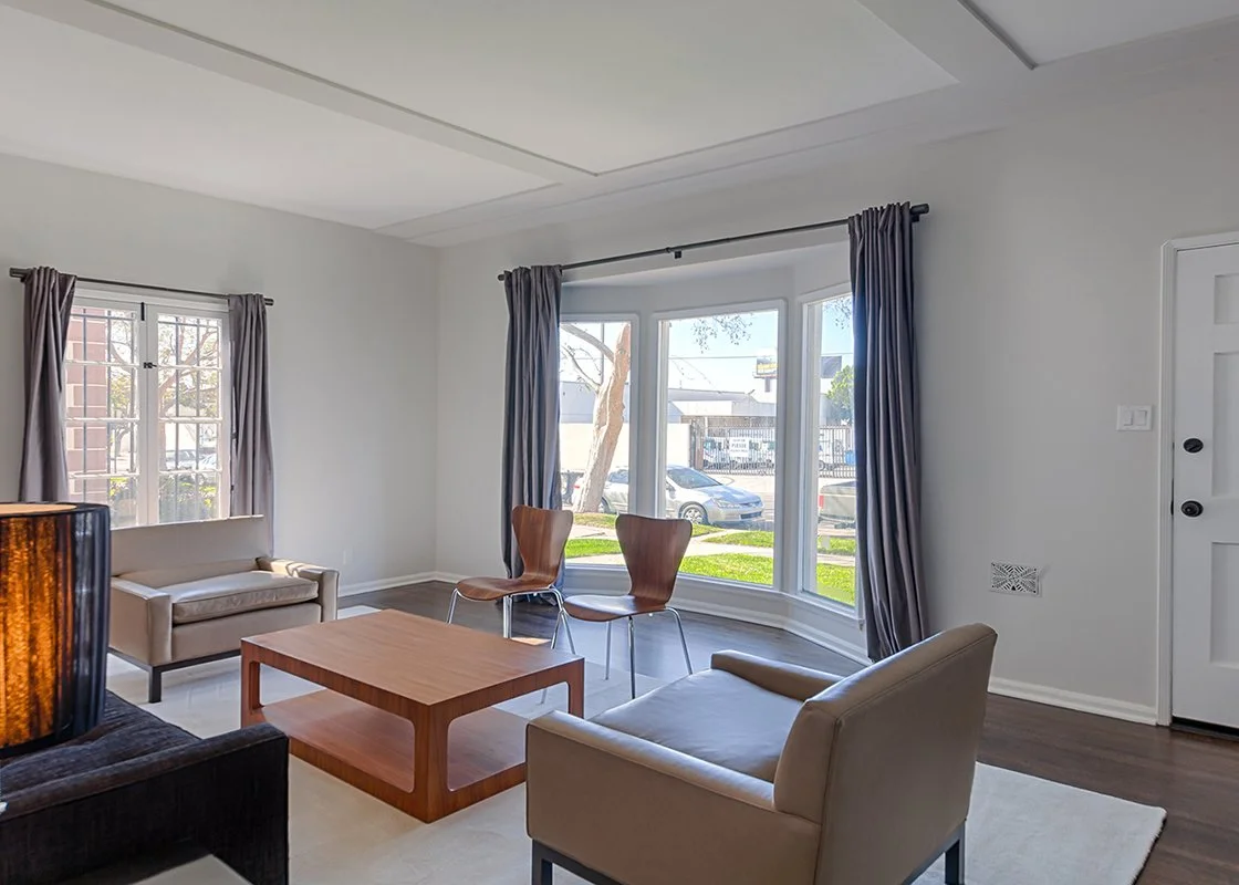 Living room with a beige sofa, two wooden chairs, a coffee table, and large windows with gray curtains, overlooking a street.