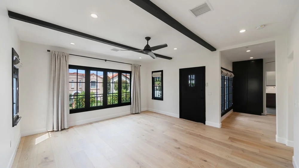 Empty living room with large black-framed windows, white walls, light wood floors, a black ceiling fan, and black accents.