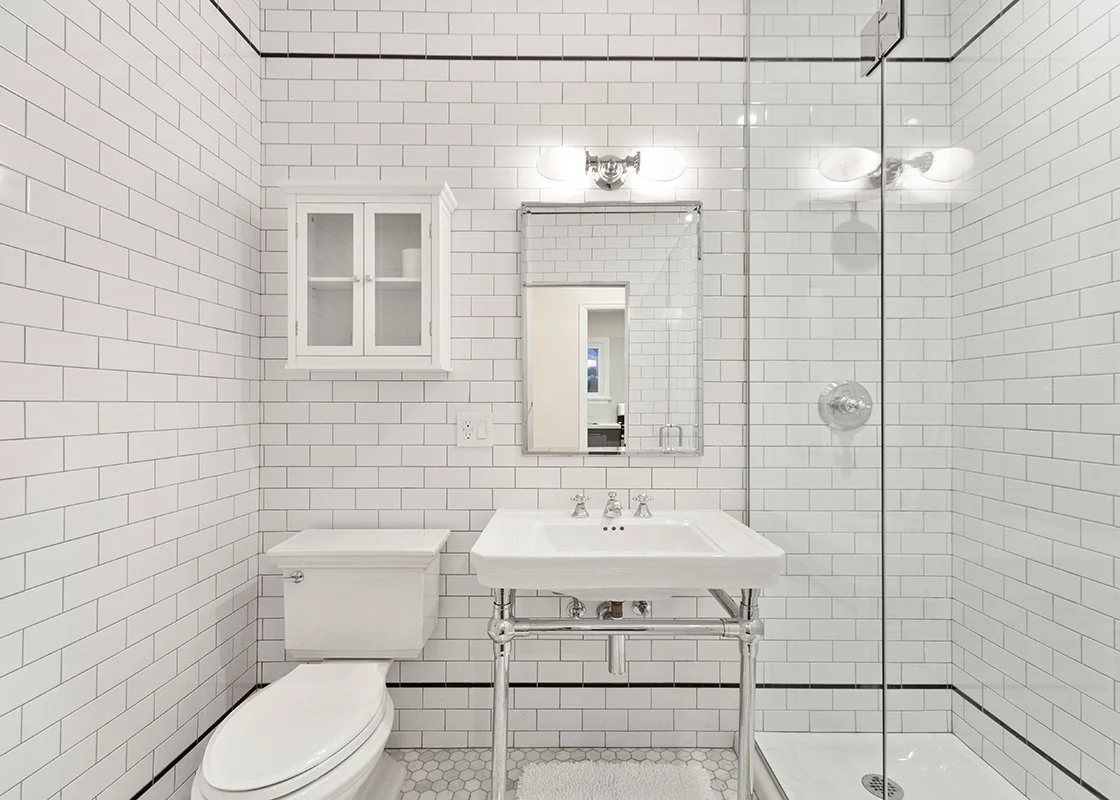 Bathroom with white subway tile walls, a toilet, a vintage-style sink, a mirror, a small wall cabinet, and a walk-in shower with a glass divider.