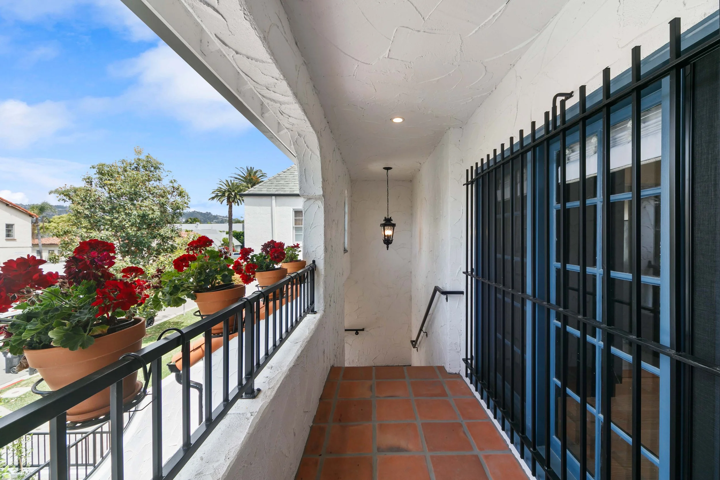 View of a balcony with terracotta tile flooring, flower pots with red flowers, black metal railing, white textured walls, black security bars on sliding glass door, hanging lantern light, and scenic background of trees and houses under a blue sky.
