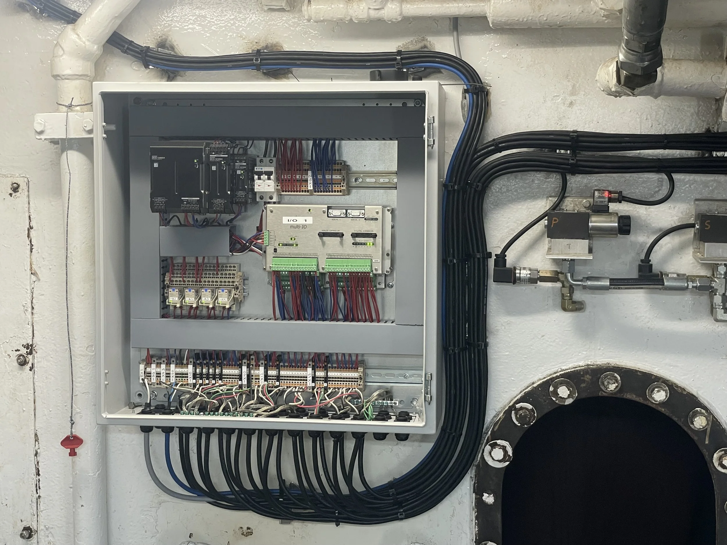 Electrical control panel with various wiring, circuit breakers, and connectors mounted inside a gray metal box, alongside pipes and fittings on a white wall.
