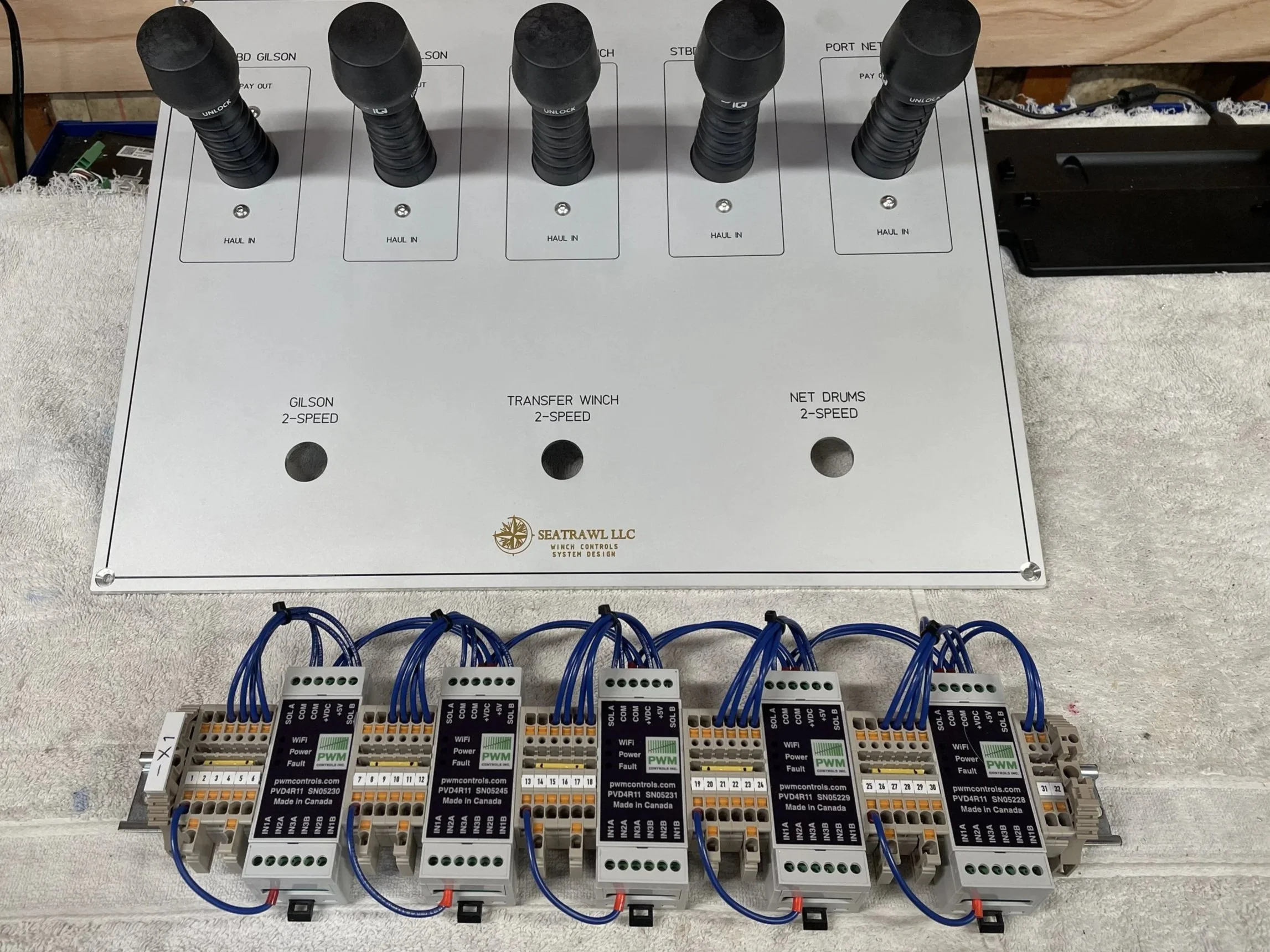 A wiring control panel with multiple terminal blocks connected to blue wires, with a white top panel labeled with control system info, and black-dialed control switches at the top labeled for winch and net drum controls.