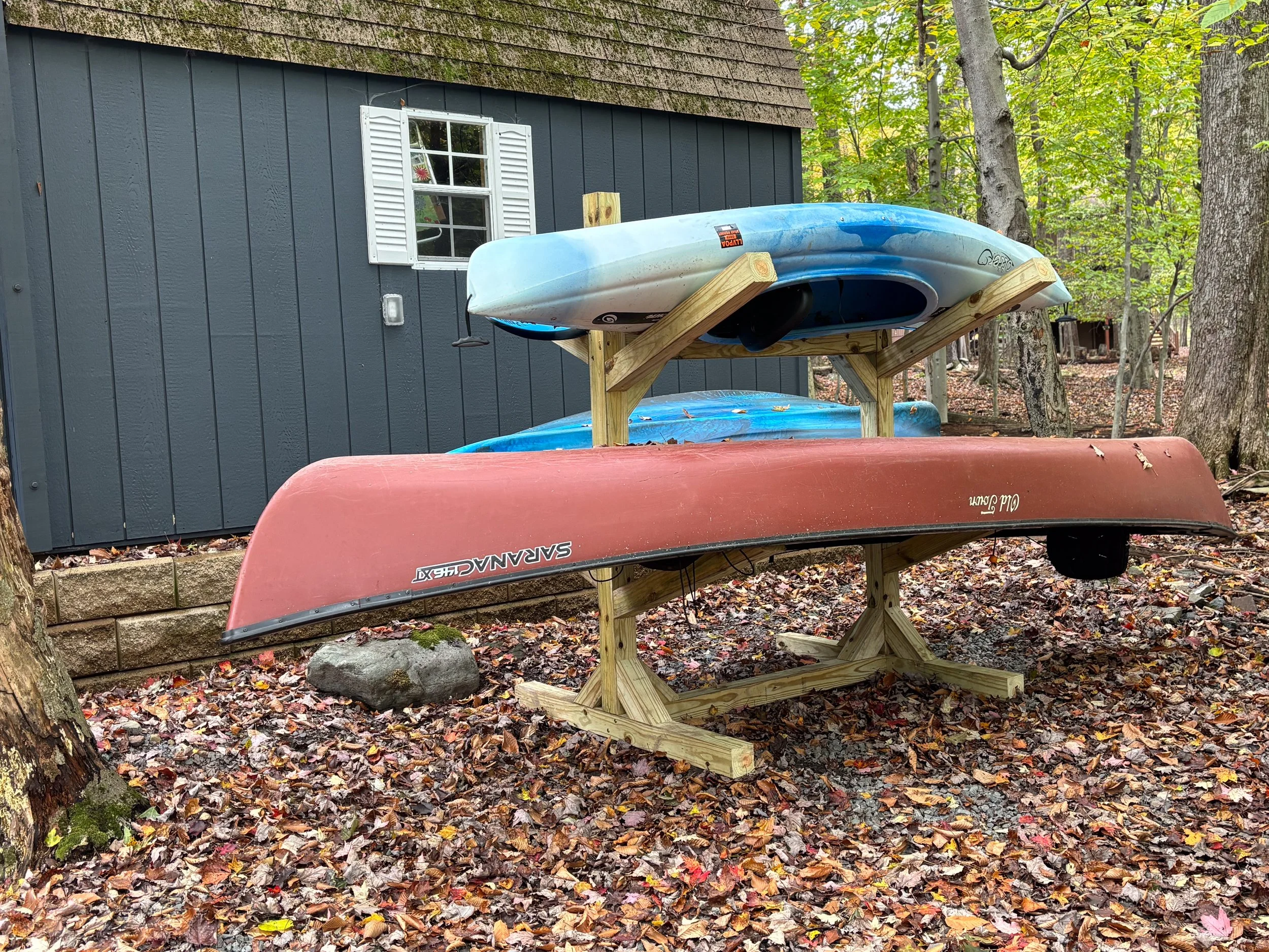 Three kayaks stacked on a wooden rack with autumn leaves on the ground, trees, and a blue house with white window shutters in the background.