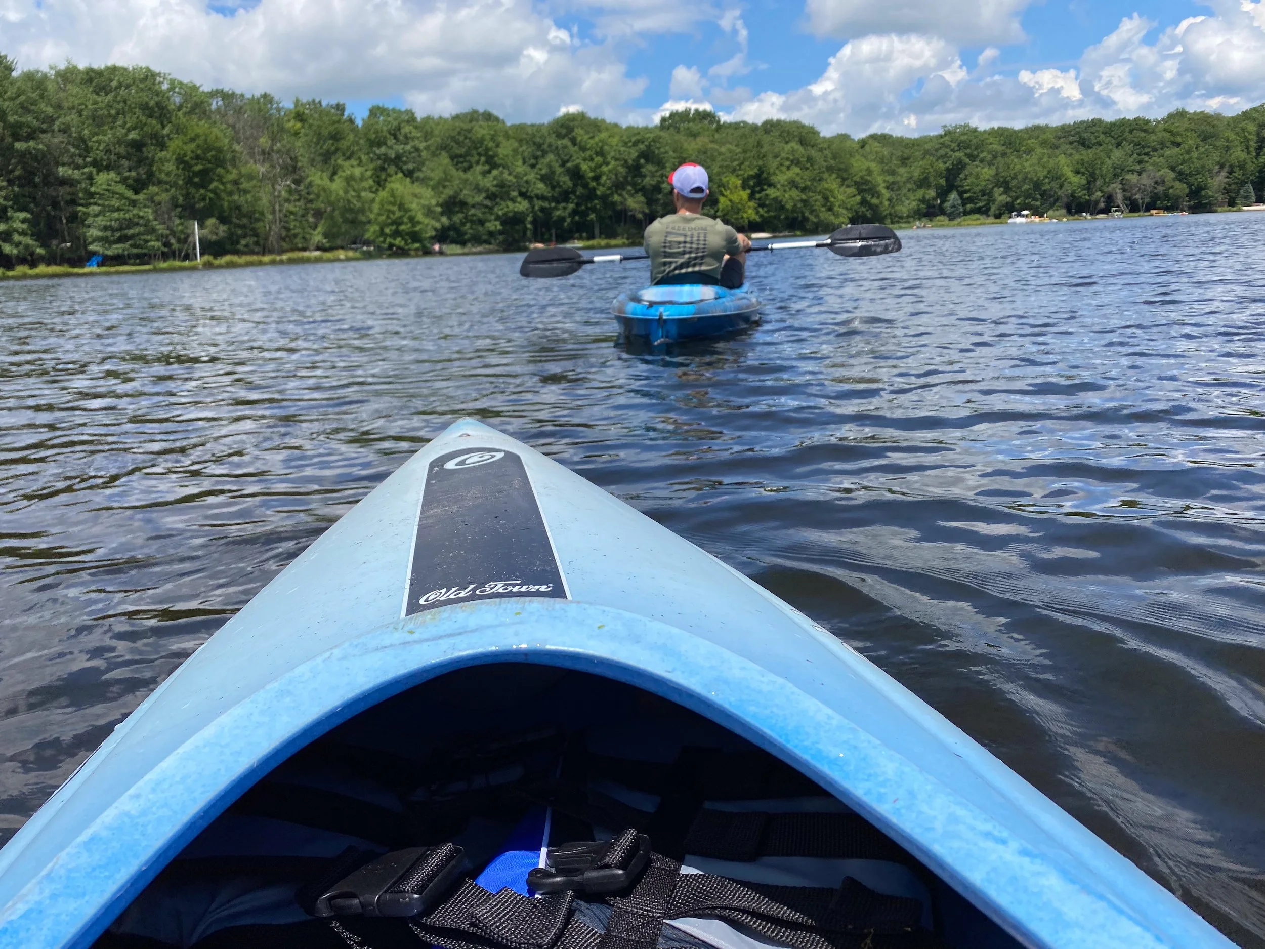 View from a kayak looking at a person kayaking on a lake surrounded by green trees under a partly cloudy sky.