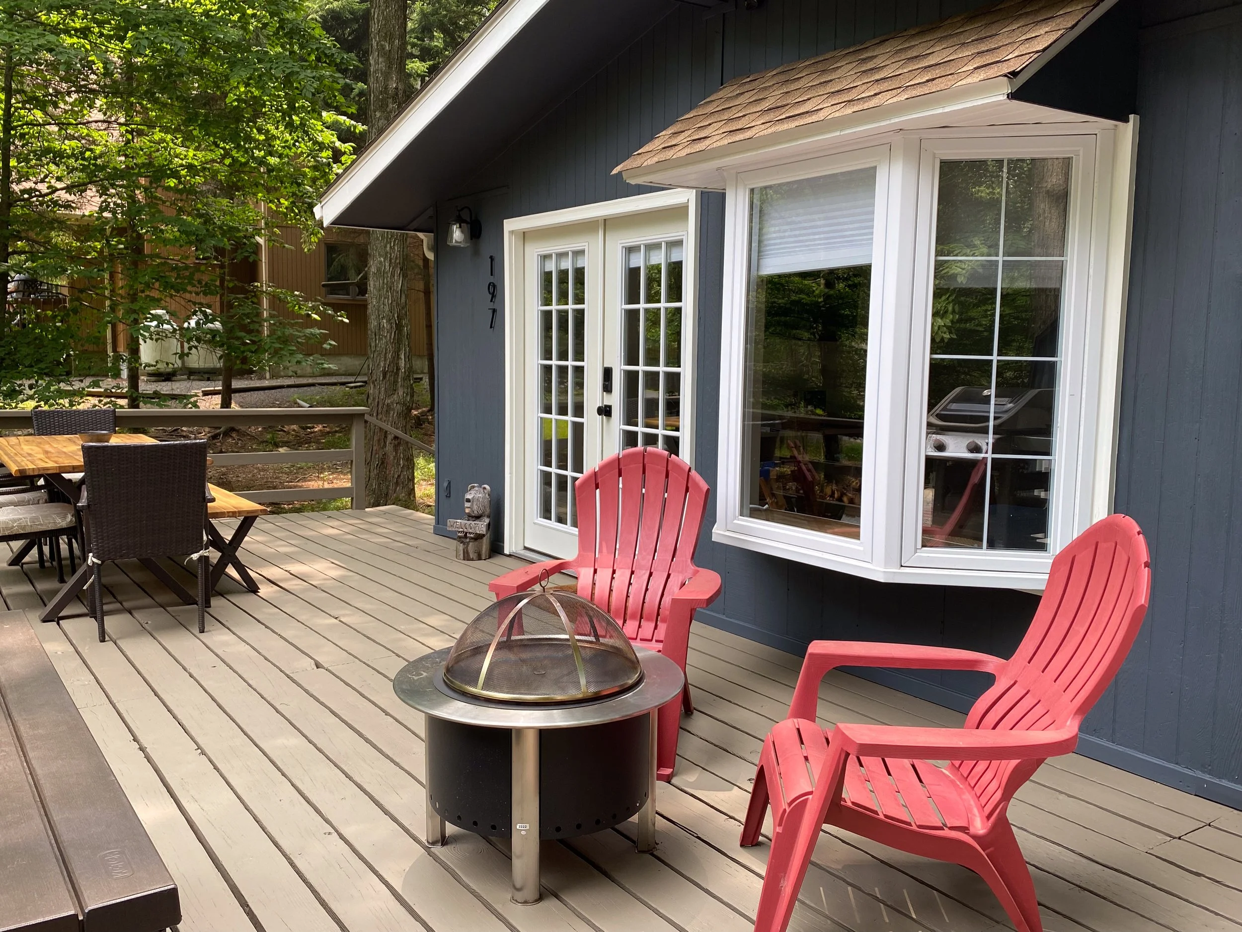 A wooden deck outside a house with a fire pit, two red Adirondack chairs, a black dining table with six chairs, and a large window on a blue house wall surrounded by a forested yard.
