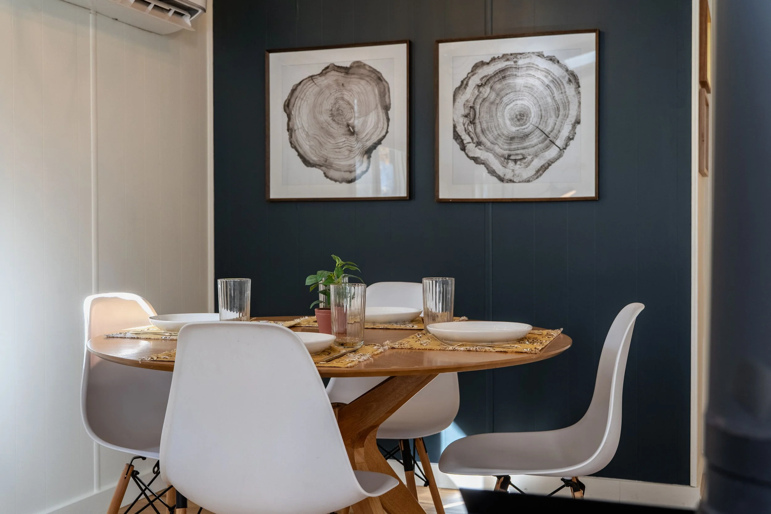 A dining room with a round wooden table set with white plates, glasses, and a centerpiece with small potted plants. Four white chairs surround the table. The wall behind features two framed black-and-white pictures of tree cross-sections. An open doo