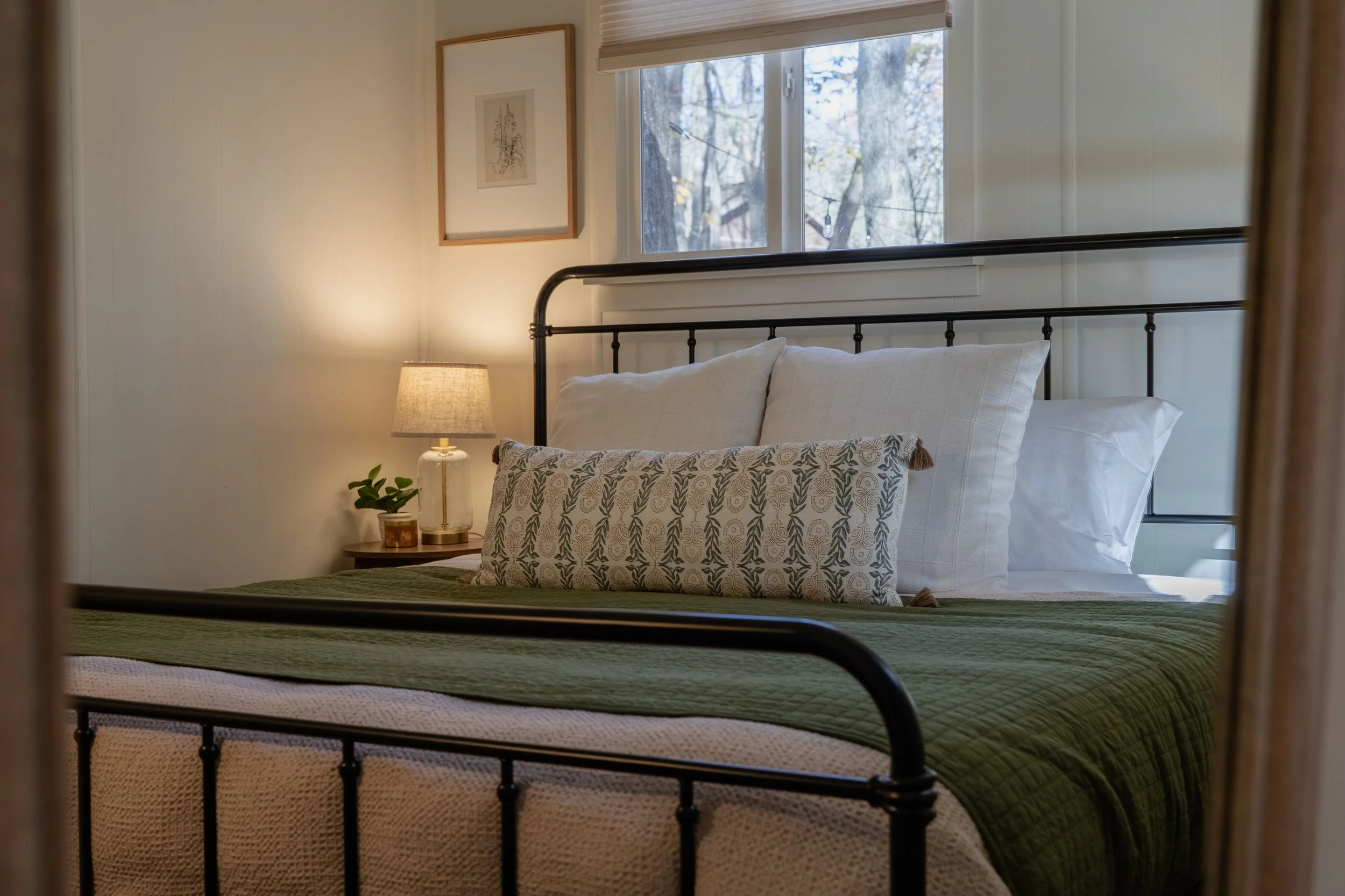 A bedroom with a king size bed,  white bedding, multiple pillows, two bedside tables with lamps, framed artwork on the walls, and windows with light filtering shades giving an outdoor view of the woods.