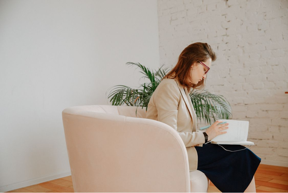 A woman with brown hair and glasses sitting in a beige armchair, reading a book in a room with white walls and a brick accent wall, with a potted plant in the background.