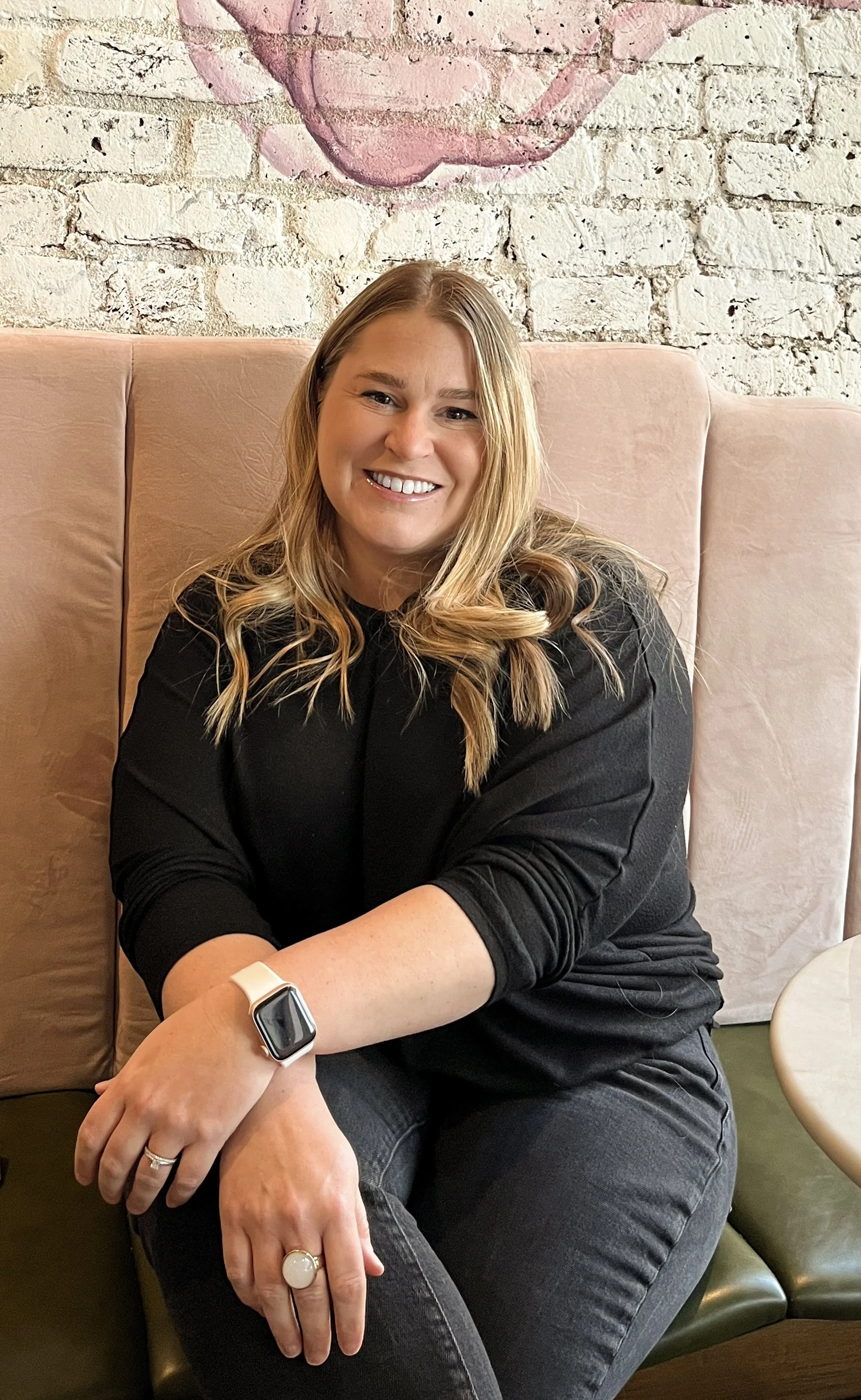 A woman with long blonde hair, smiling, sitting on a pink booth with a white brick wall behind her. She is wearing a black long-sleeve top, a smartwatch, and rings on her fingers. There is a painted face on the wall above her.