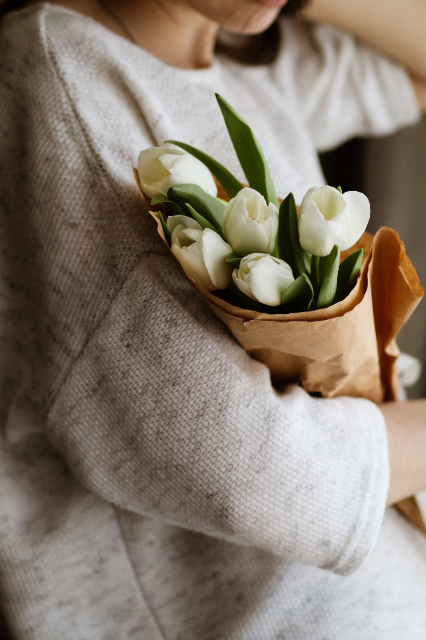 Person in a white sweater holding a bouquet of white tulips wrapped in brown paper.